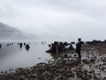 Full length side view of woman standing by river against sky during foggy weather