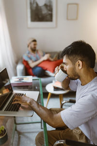 Side view of man drinking coffee while using laptop at home