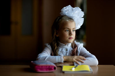 Thoughtful girl sitting with book and pouch at desk in school