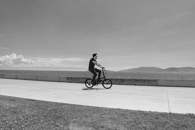 Side view of young man riding bicycle on road by sea against sky during sunny day