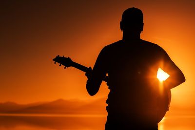 Silhouette man playing guitar against orange sky during sunset