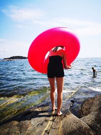 Rear view of girl standing with pink inflatable ring at beach against sky