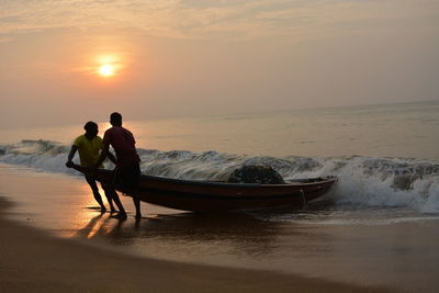 Fishermen pulling boat at beach against sky during sunset