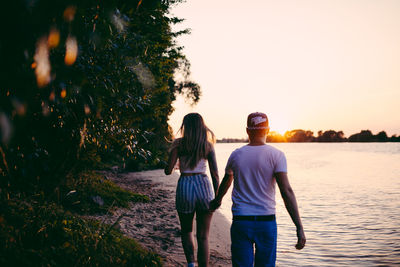 Rear view of couple holding hands while walking at beach against sky