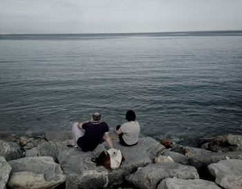 Rear view of couple sitting on rock by sea against sky