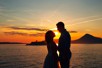 Side view of couple embracing while standing at beach at sunset