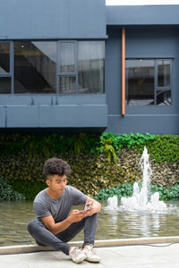 Full length of young man sitting on fountain against building