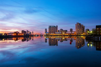 Illuminated buildings by river against sky at sunset