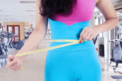 Midsection of woman measuring stomach while standing at gym