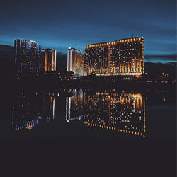 Illuminated modern buildings by river against sky at night