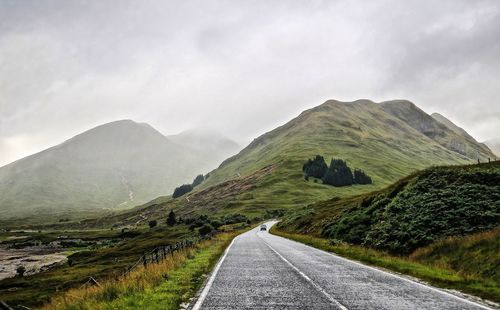 Road leading towards grassy mountains against cloudy sky
