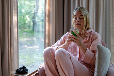 A blond woman in pink pajama sits in the pink armchair and sips mint tea.
