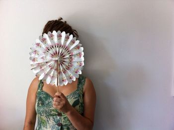 Woman hiding face with hand fan against white wall