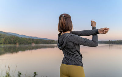 Back of a beautiful young asian woman in warm up jacket stretching her arms up in front of a lake