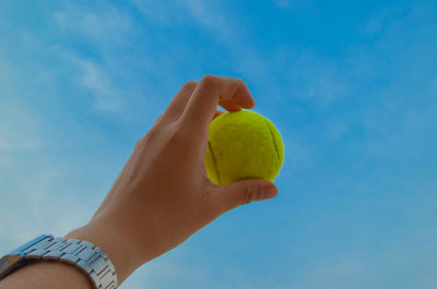 Cropped hand of person holding tennis ball against blue sky