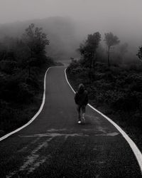 Rear view of man walking on road amidst tees during foggy weather