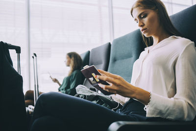 Young businesswoman looking at passport while sitting in airport departure area