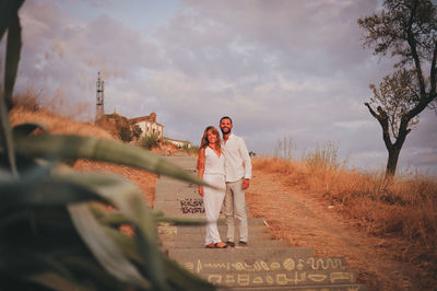 Full length of young couple standing on steps against cloudy sky