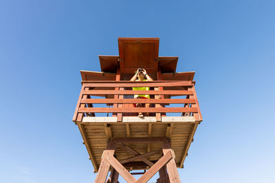 Low angle of looking through binoculars on wooden watchtower while overseeing safety at sea against cloudless blue sky