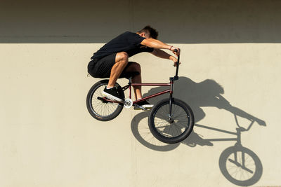 Full body side view male in casual clothes jumping on bmx bicycle against sunlit concrete wall on summer weekend day on street