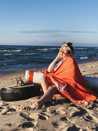 Young woman with orange shawl by tire at beach against sky