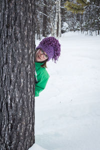 Cute little girl wearing glasses and looking out from behind a tree in forest during winter