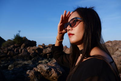 Woman shielding eyes while standing by rock against sky