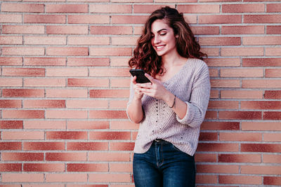 Beautiful young woman using mobile phone against brick wall