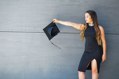 Woman holding mortarboard while standing against wall