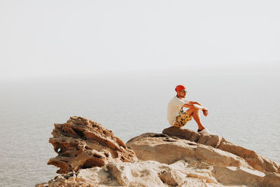 Man sitting on rock by sea against clear sky