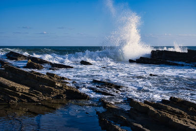 Waves splashing on rocks at shore against sky