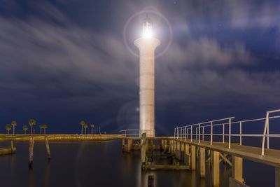 Low angle view of lighthouse by pier over sea against cloudy sky at dusk