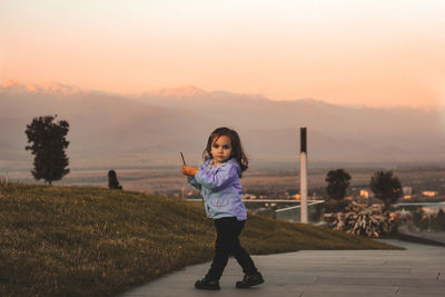 Portrait of cute girl standing on steps in park against sky during sunset