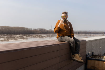 Full length of man drinking smoothie sitting on wall against sky