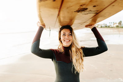 Smiling surfer woman dressed in wetsuit standing looking at camera while carrying surfboard above head on the beach during sunrise in the background