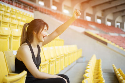 Side view of young woman cheering while sitting at stadium