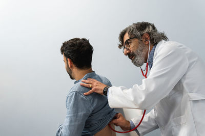 Side view of elderly male doctor examining patient with stethoscope during appointment in hospital against white wall