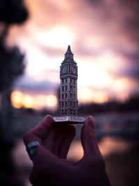 Cropped hand of person holding big ben miniature against sky during sunset