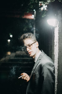 Portrait of businessman smoking cigarette by illuminated wall at night