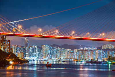 Illuminated bridge over river by buildings against sky at night