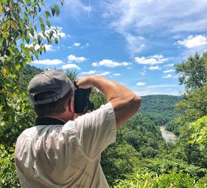 Rear view of senior man photographing with camera while standing on mountain against blue sky during sunny day