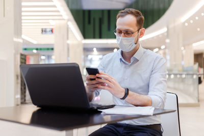Businessman wearing mask using phone while sitting at mall