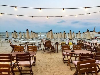 Empty chairs and tables at beach against sky