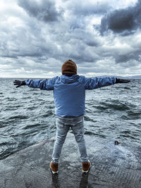 Rear view of man with arms outstretched standing by sea against cloudy sky