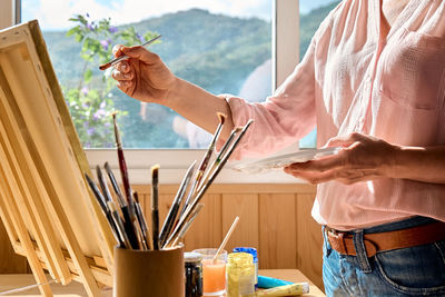 Female hand holding a paintbrush near canvas on easel near the window in the studio.