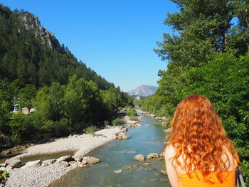 Rear view of redhead woman looking at river in forest against clear sky