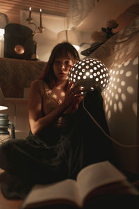 Young woman sitting in illuminated electric lamp at home