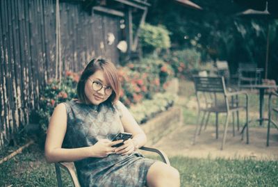 Portrait of young woman using phone while sitting on chair in lawn
