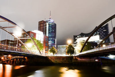 Illuminated bridge over river by buildings against sky at night