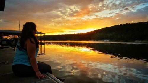 Woman sitting by lake against sky during sunset